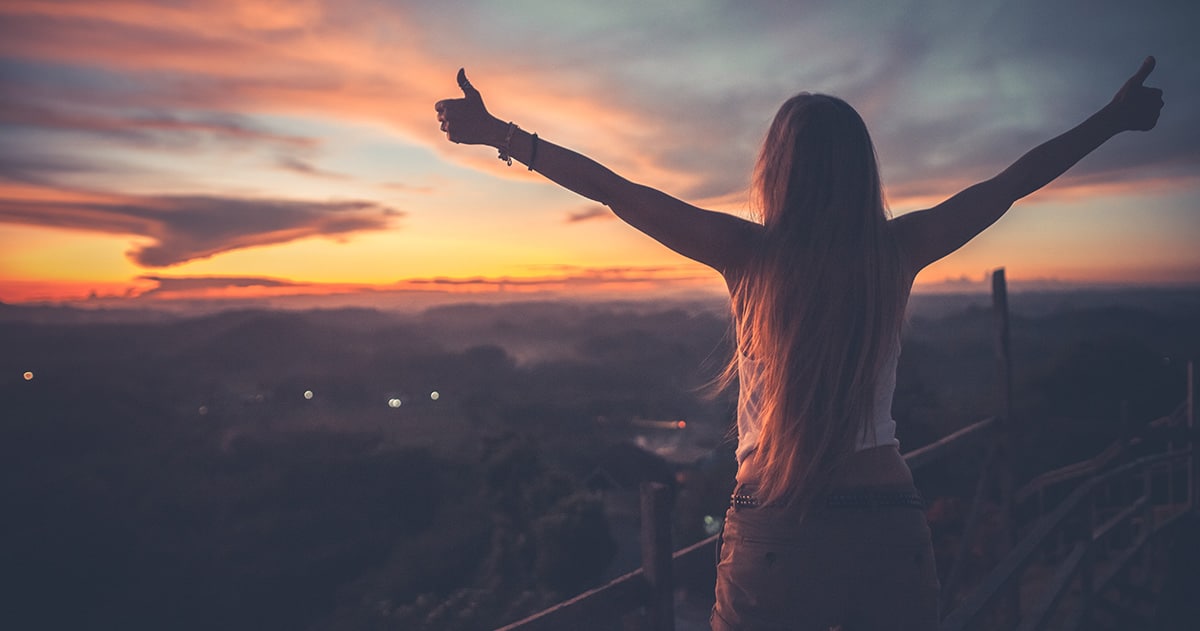 Woman standing on edge of cliff with arms spread and thumbs up.